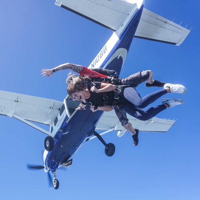 Two people skydive in tandem from a blue and white airplane against a clear blue sky. They appear excited and are in free fall with parachute harnesses. The aircraft is visible above them, and one engine propeller can be seen.