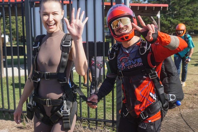 A smiling woman in a brown skydiving suit waves to the camera, while a person in a red skydiving outfit and helmet gives a peace sign. They are outdoors, near a black metal fence and green trees in the background.