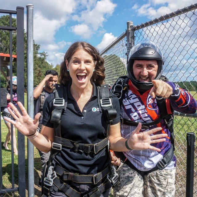 A woman and a man in skydiving gear stand near a fence, smiling and gesturing enthusiastically. The woman has her hands raised in excitement, while the man points towards the camera. The background shows a grassy area and a partly cloudy sky.