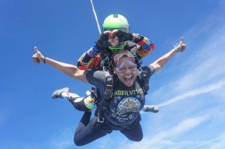 A woman and an instructor tandem skydiving. The woman is smiling broadly, giving a thumbs-up, while wearing a black shirt. The instructor behind her wears colorful gear and a green helmet. The background is a clear blue sky.