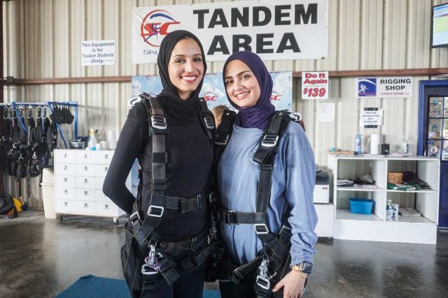 Two people wearing hijabs and skydiving harnesses stand smiling in a tandem skydiving area. They are in a room with skydiving equipment visible in the background.