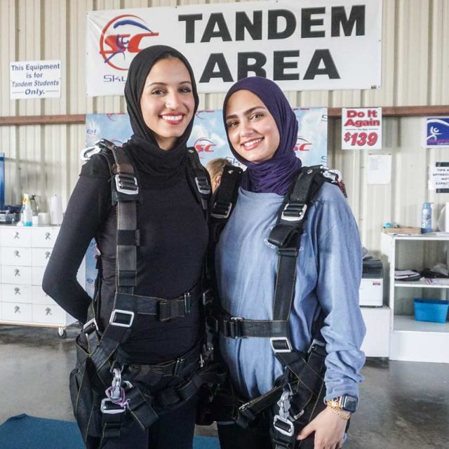 Two people wearing hijabs and skydiving harnesses stand smiling in a tandem skydiving area. They are in a room with skydiving equipment visible in the background.